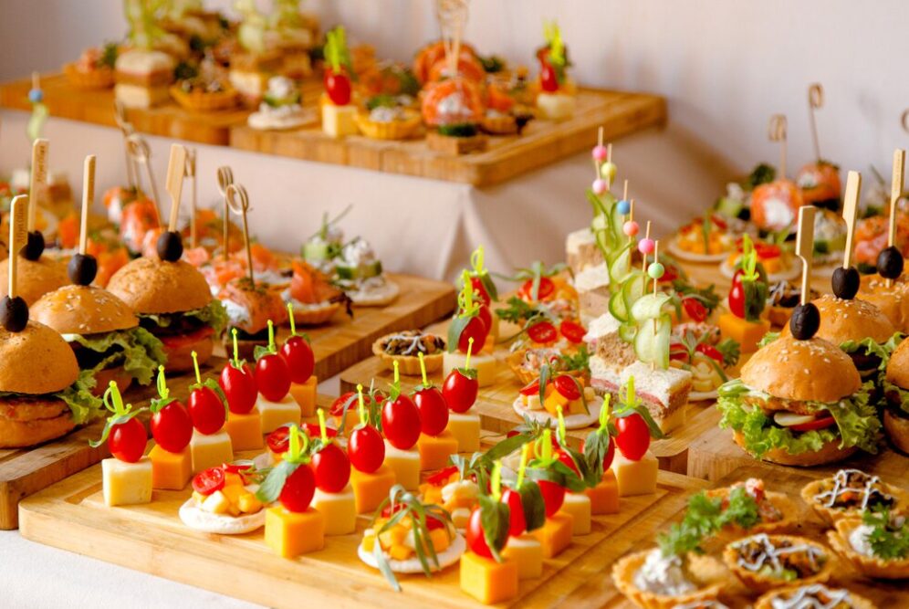 Assortiment coloré d'amuse-bouches comprenant des mini-burgers, brochettes de fromage et tomates cerises, canapés et légumes frais, présenté sur des plateaux en bois, parfait pour un service traiteur.